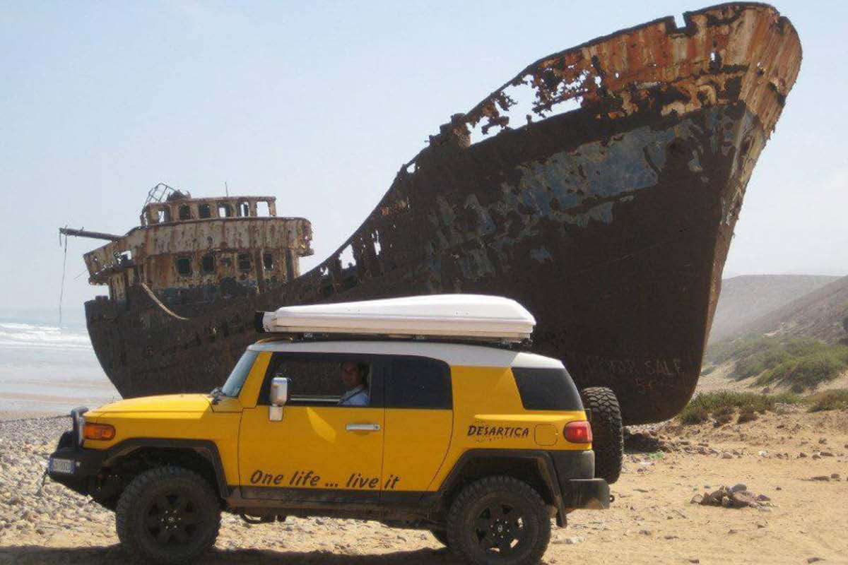 Das bild zeigt einen gelben Geländewagen vor einem Schffswrack